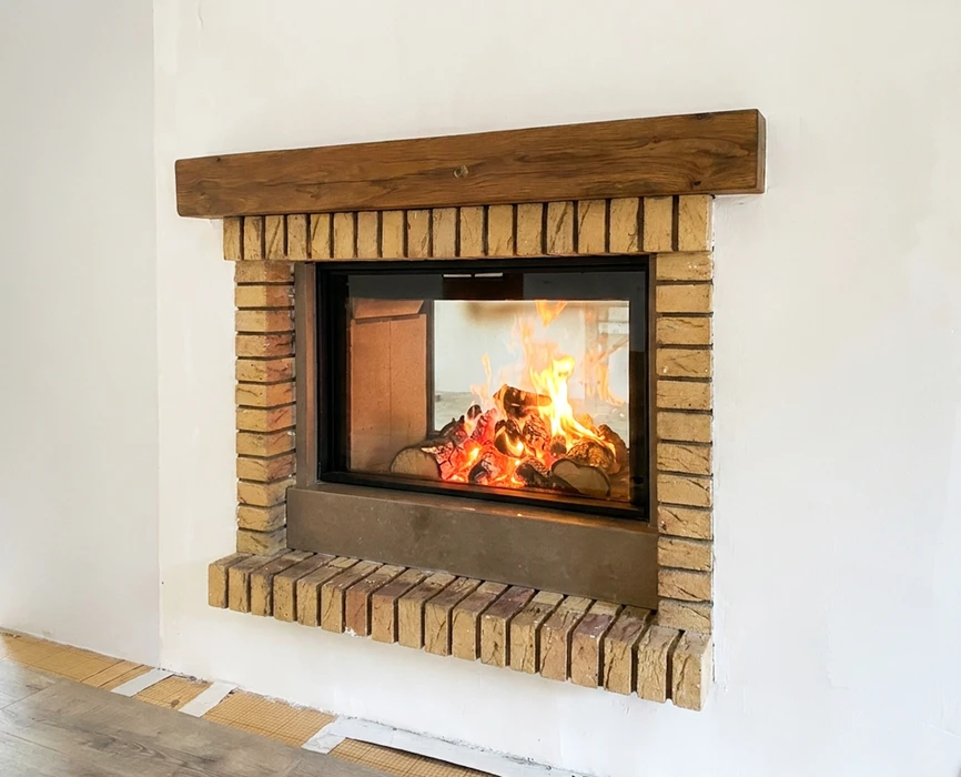 Cheminée encastrée à foyer fermé, encadrée de briques de parement beige et d'un manteau en poutre de bois massif