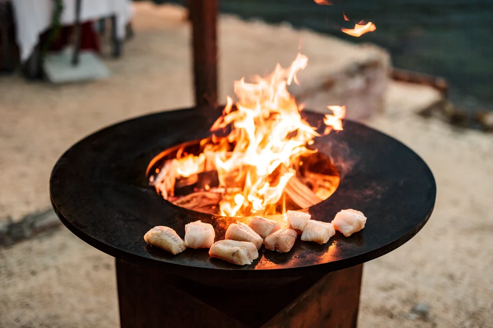Scène extérieure avec brasero plancha circulaire en métal noir sur pied, flammes vives au centre, morceaux de viande ou légumes roulés disposés sur la surface de cuisson chaude autour du foyer