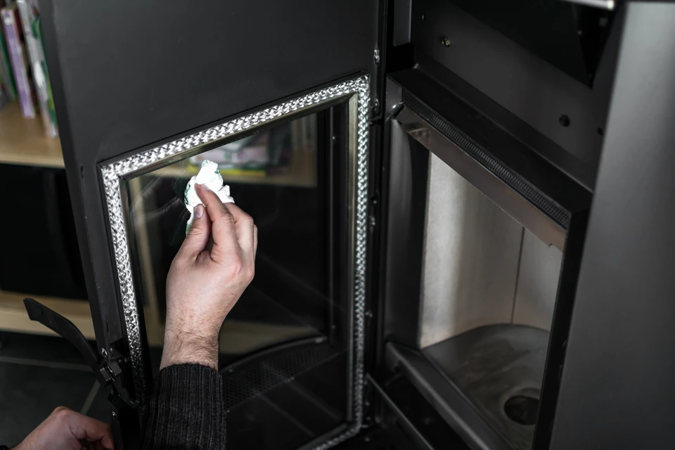 Une personne nettoie la vitre d'un foyer de cheminée avec un chiffon blanc dans un intérieur résidentiel