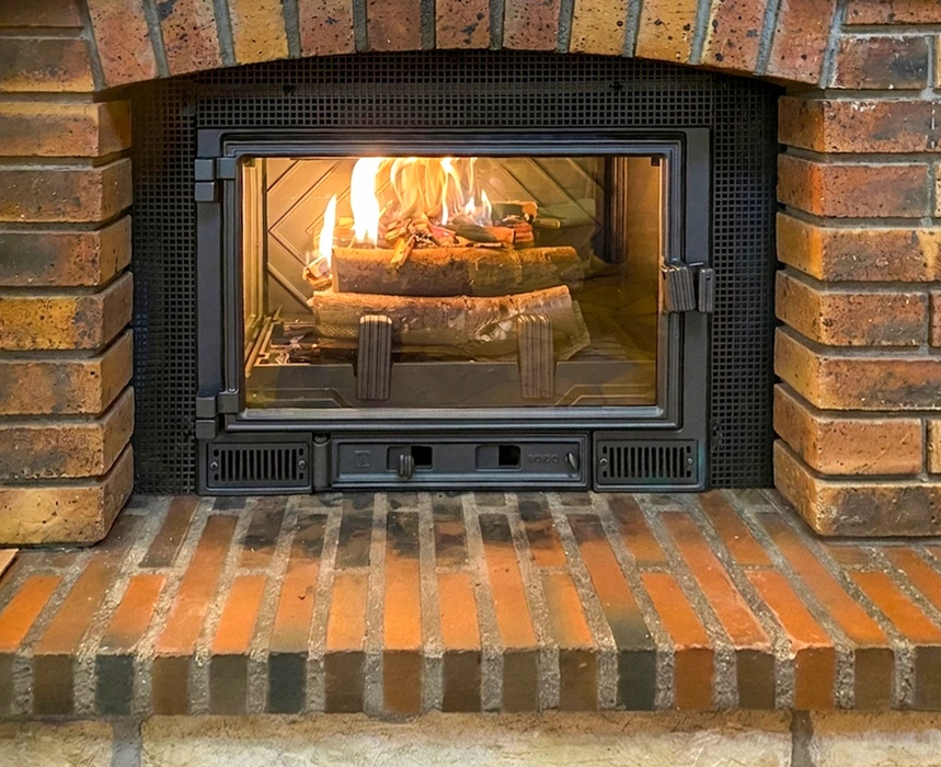 Cheminée intérieure en briques rouges avec insert à bûches vitré, cadre métallique noir avec joints coupe-feu, grilles de ventilation basse, poignée centrale et tiroir de régulation d'air